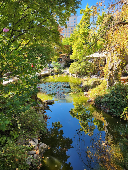 A photo from the Lan Su Chinese Garden in Portland. A pond is surrounded by trees, which are reflected in the still water. The photo is oddly symmetrical, almost unsettling in that it's difficult to tell what is a reflection and what is the actual vegetation. In the background, you can tell that it's an urban garden, as there's a large office building (also partially reflected in the pond).