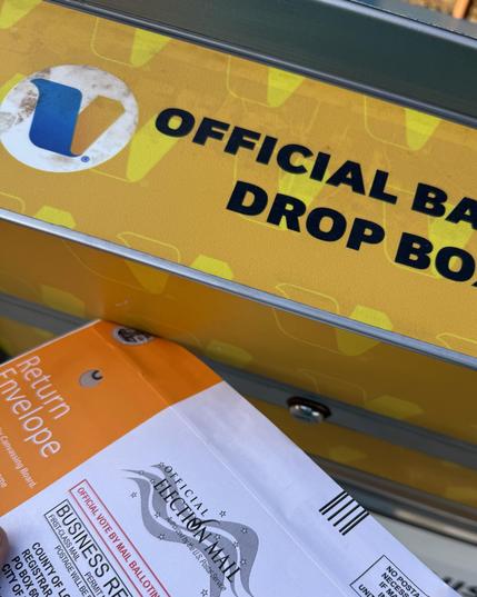 A closeup of an official ballot drop box with an orange return envelope, alongside a label stating "OFFICIAL BALLOT DROP BOX."
