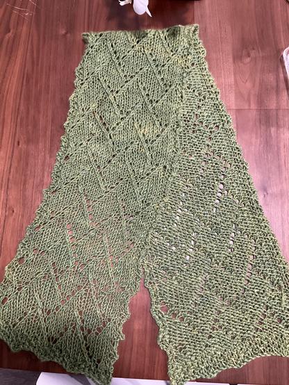 A green scarf folded in half on a wooden table. The halves are spread out so you can see both sides of the knitting pattern.
