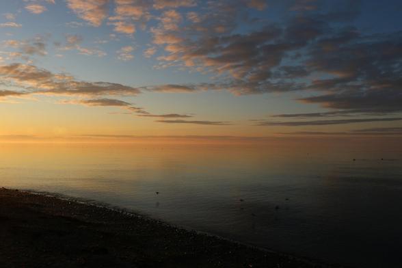 Photograph of a sunset over the sea, in shades of orange and blue, with gray clouds illuminated by orange light. A few seabirds are flying over the water and gulls glide on the almost calm surface, reflecting the light and colors of the setting sun. The sand and stone shore is visible in the lower left corner.

Photographie d'un coucher de soleil sur la mer, dans des tons dégradés de orange et de bleu , avec des nuages gris éclairés par la lumière orange. Quelques oiseaux marins sont en vol au-dessus de l'eau et des goélands glissent à la surface presque calme, sur laquelle se reflète la lumière et les couleurs du soleil couchant. Le rivage de sable et de pierre est visible dans le coin inférieur gauche.