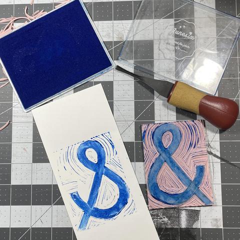 Block printed ampersand carved from pink rubber in blue ink on white paper. The ampersand is carved forward and printed backwards, due to distraction.