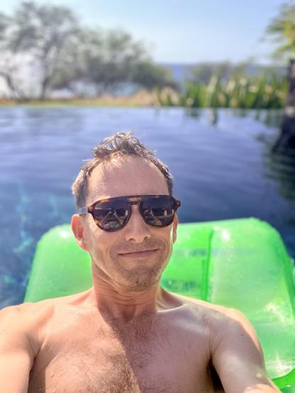 Selfie of an old man on a pool floaty in an infinity pool off the ocean. The selfie is cropped just above his nipples.