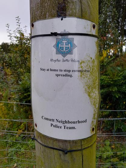 Laminated paper notice pinned to a telegraph pole. Wire fence and trees behind, rural setting. The notice has been attached with cable ties and drawing pins which have gone rusty. Algae are growing up one side.

"Durham Constabulary [logo]
Together Better Policing [cursive]

Stay at home to stop coronavirus spreading.

Consett Neighborhood Police Team."