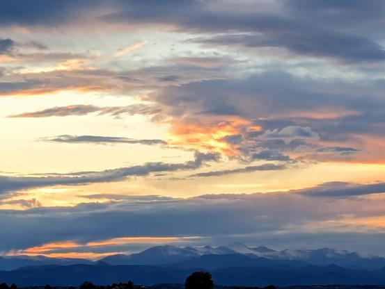 A mountain range at dusk with diffused light lighting up orange clouds constrasting against the blue tones of the silhouette of the snowcapped peaks.