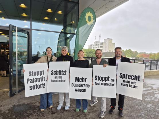 5 Menschen mit Plakaten vor eine Beachflag der Grünen und dem Messegebäude Offenburg:
Stoppt Palantir, sofort!
Schützt unsere Grundrechte! 
Mehr Demokratie wagen!
Stoppt die Tech-Oligarchie!
Sprecht mehr mit eurer Basis!
