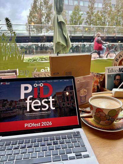 Un ordinateur portable posé sur une table de café affiche le site web de PIDfest, avec le logo PIDfest sur une photo d’un canal de Leyde et une bannière rouge indiquant « PIDfest 2026 ». À côté se trouve un cappuccino dans une tasse décorée de boutons colorés. Derrière l’ordinateur, on aperçoit des magazines et des menus ; à travers la grande vitre du café, un cycliste passe le long d’une rambarde par une journée nuageuse, avec des arbres et un bâtiment en arrière-plan.