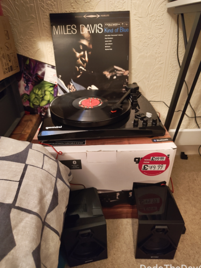 a record player by Gemini, sitting on its box, the two speakers on the floor, a record is playing and the sleeve propped up against the wall behind it. this is all squeezed in between my desk, bed and TV.