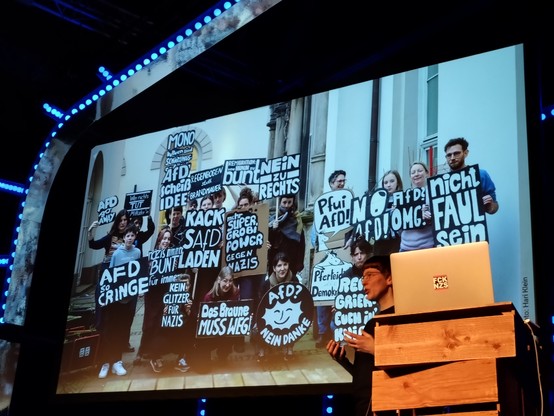 Chris standing in front of a display, showing a photo of lots of people who took her workshop and holding fantastic and punchy monochrome signs that they made.