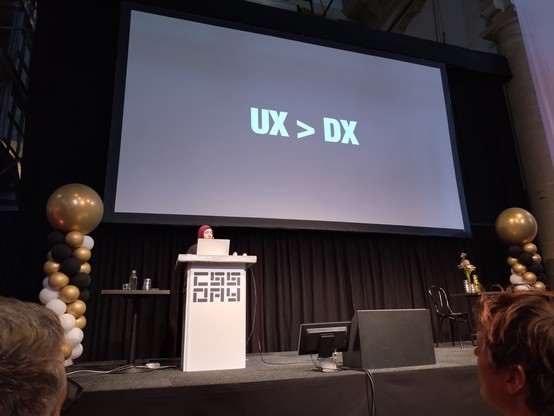 Sara behind podium with bolded text "UX > DX" alone on the screen behind her.