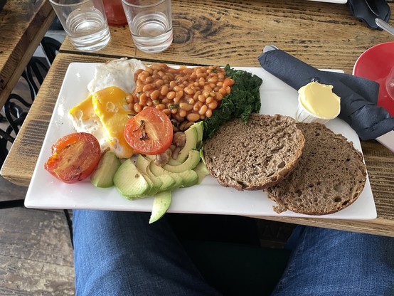 Photo of long white rectangular plate, covered in two bread rolls, baked beans, fried tomatoes, fried eggs, avocado, and spinach. It was /very/ tasty!