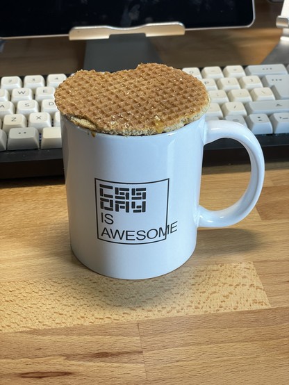 A photo of a mug in front of a computer keyboard. The mug is white, and has “CSS Day is awesome” printed on it. There is a Dutch stroopwafel balanced on top of the mug.