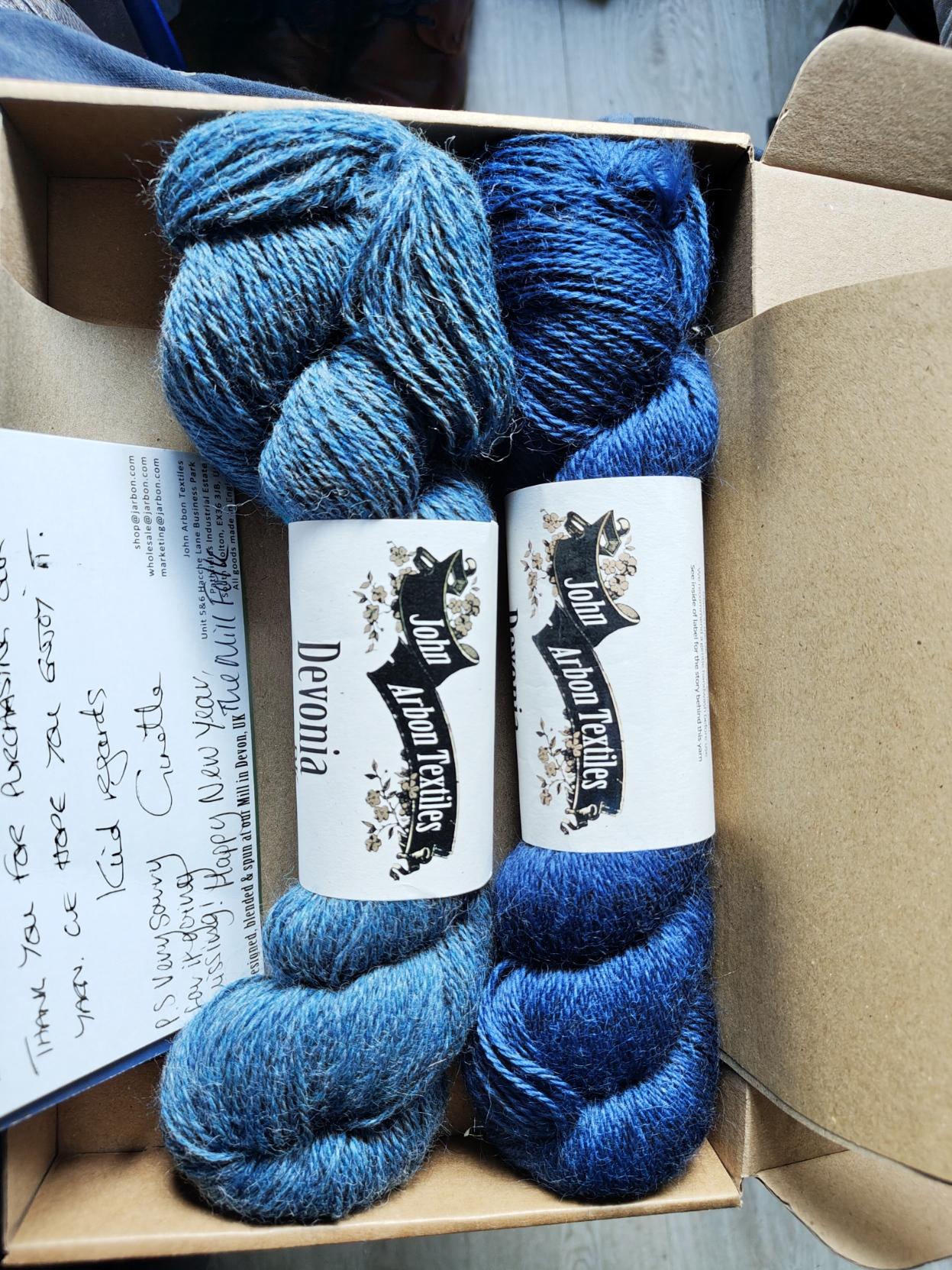 Two skeins of yarn. Both a blue, one dark one lighter. Both have a band with John Arbon Textiles, the model is Devonia DK.
They are both sitting in the shipping box and a note can be seen on the left hand side, hand written saying thank you for your purchase.