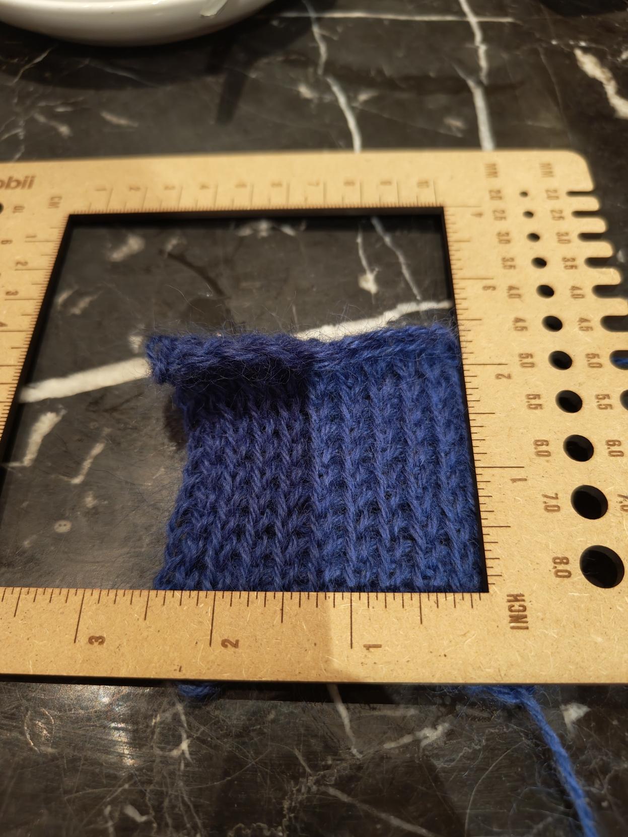 a blue yarn tension square on a black marbled table with a ruler overlay 