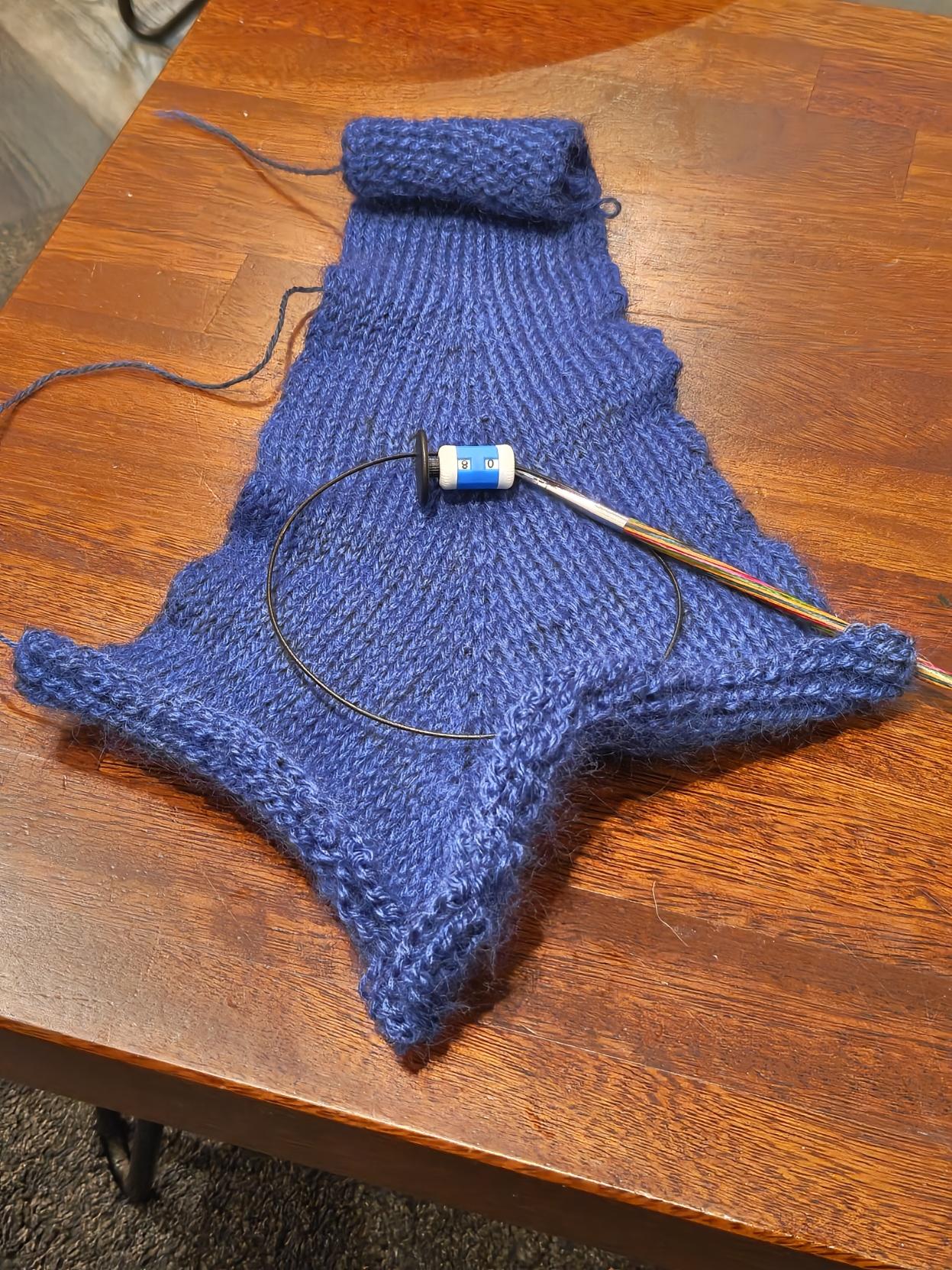 A woollen swatch in blue on a wooden table. It tapers out from 3 inches to approximately 10 inches wide and is about 15 inches long (although the edges are curling in) 