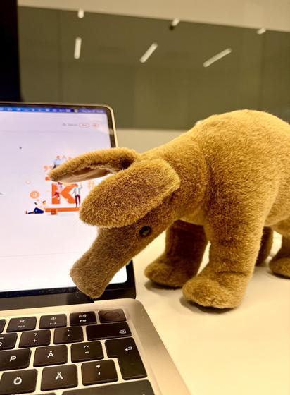 A golden-brown plush aardvark named “Muurahaiskarhu” stands on a laptop keyboard, its long snout extending toward the screen which displays the Kesko logo in orange and white. The soft toy is photographed against a minimal office setting with white pendant lights visible in the blurred background.