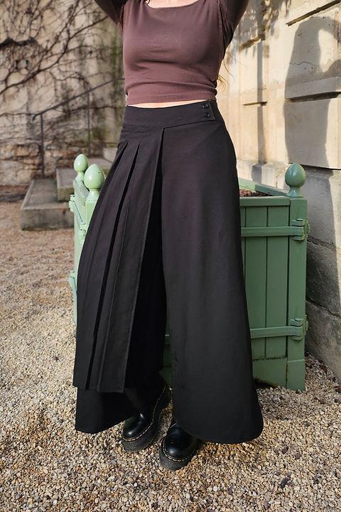 Wide leg black Japanese inspired pants with a wide pleated front panel resembling a half skirt buttoned at the waistband, made of fabric with subtle sheen, extending to the ankles.
