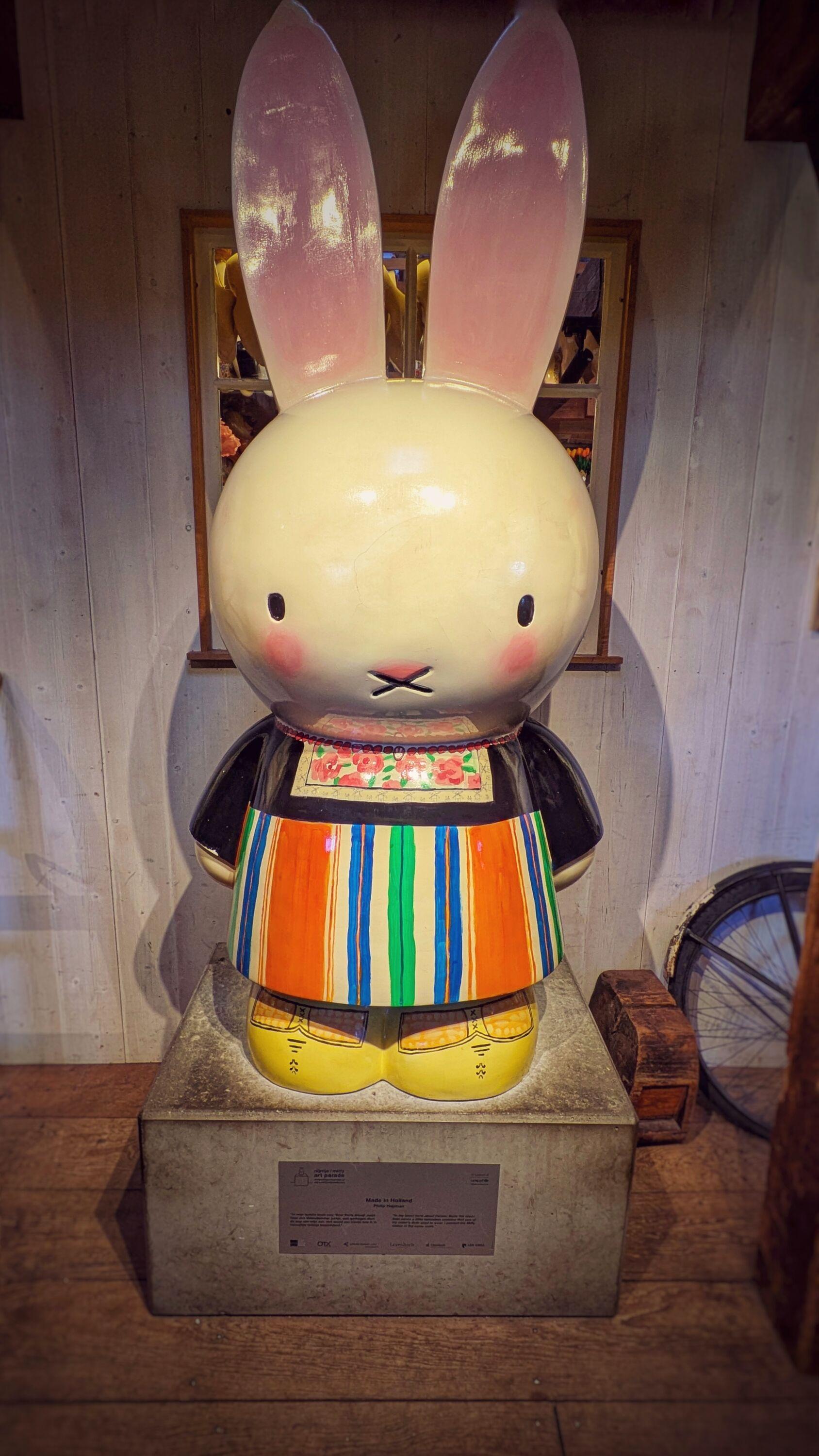 A giant wooden sculpture of Miffy (a large white bunny) dressed in traditional Dutch clothes with a colorful floral chest patch and striped skirt, and the traditional yellow wooden sandals.