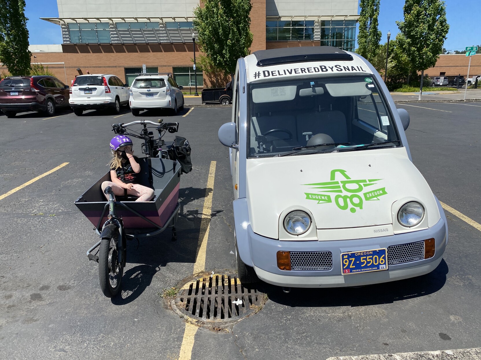 A Cetma Largo cargo bike parked next to a Nissan S-Cargo, viewed from the front, the S-Cargo says “delivered by snail.” The child in the bike is still bored.