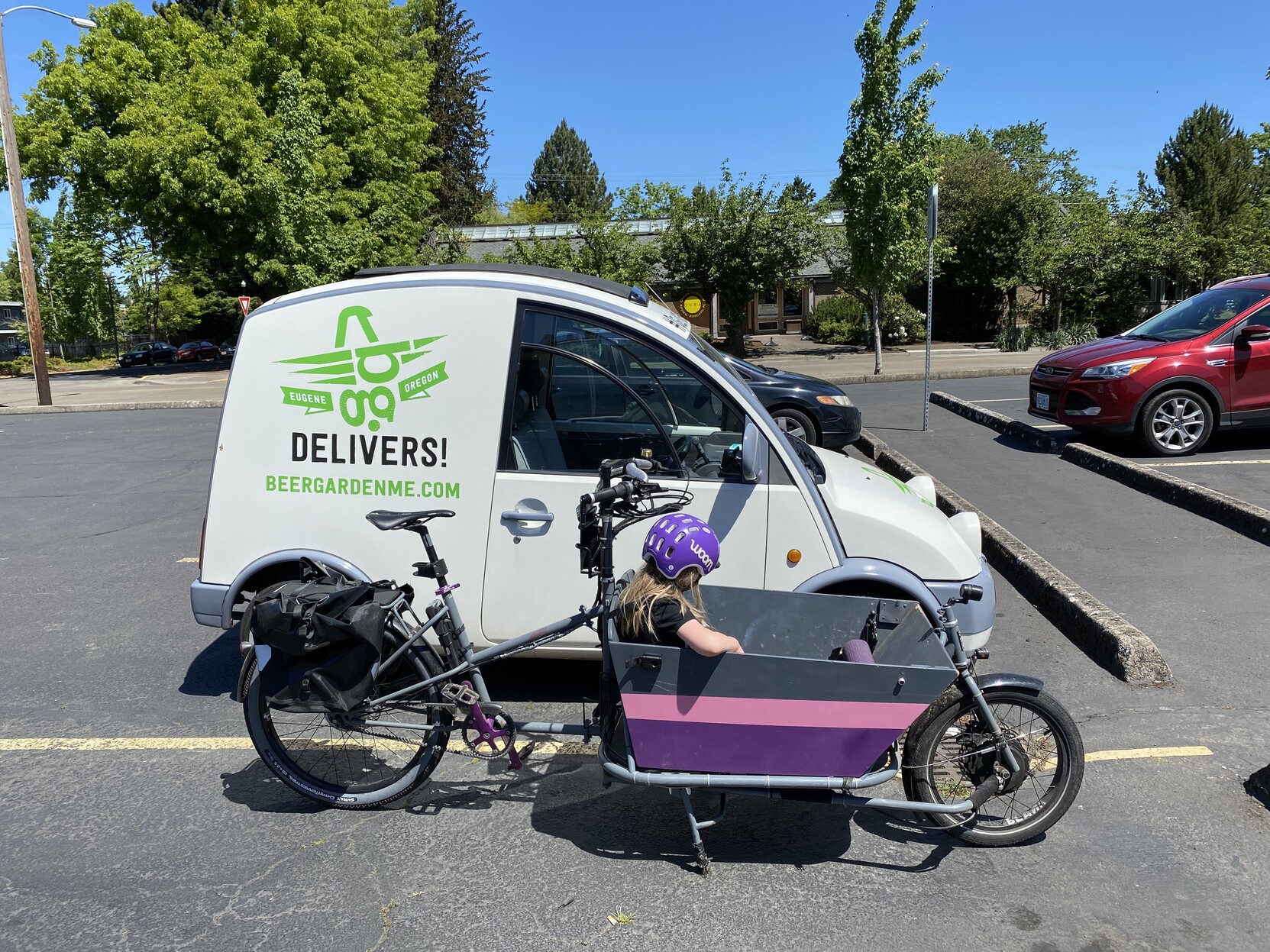 A Cetma Largo cargo bike parked next to a Nissan S-Cargo, viewed from the side showing that they’re roughly the same length. The child in the bike is bored.