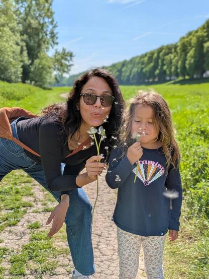 A woman and a girl blowing grass with seeds flying in the first plan. Green grass and trees behind them with plenty of open space. Blue sky above. 
The scene is intensely illuminated. 