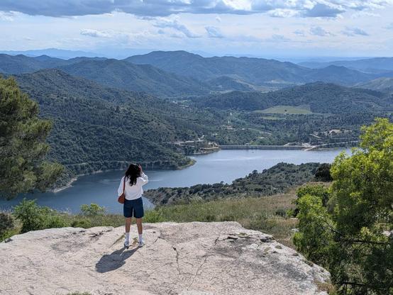 A woman standing in a rocky balcony overlooking a lake with mountains in the background. 