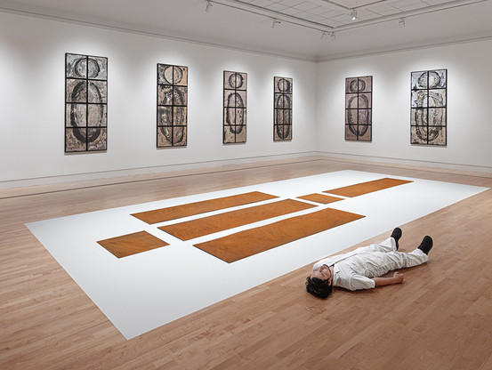 A photograph of artist Kei Ito lying on the ground in a gallery of his work, next to his installation of rusted metal panels based on the atomic bombs dropped on Japan