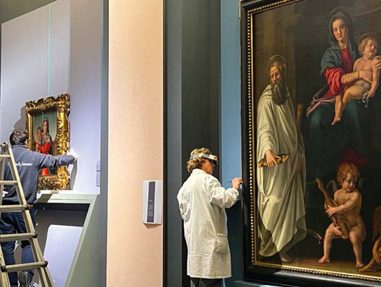 Museum art handlers and preparators putting together the final details of an exhibition by the Florentine Renaissance painter Pier Francesco Foschi. One man hangs a Madonna and Child painting in a gold frame. Another seems to be touching up wall paint behind a large painting of the holy family.