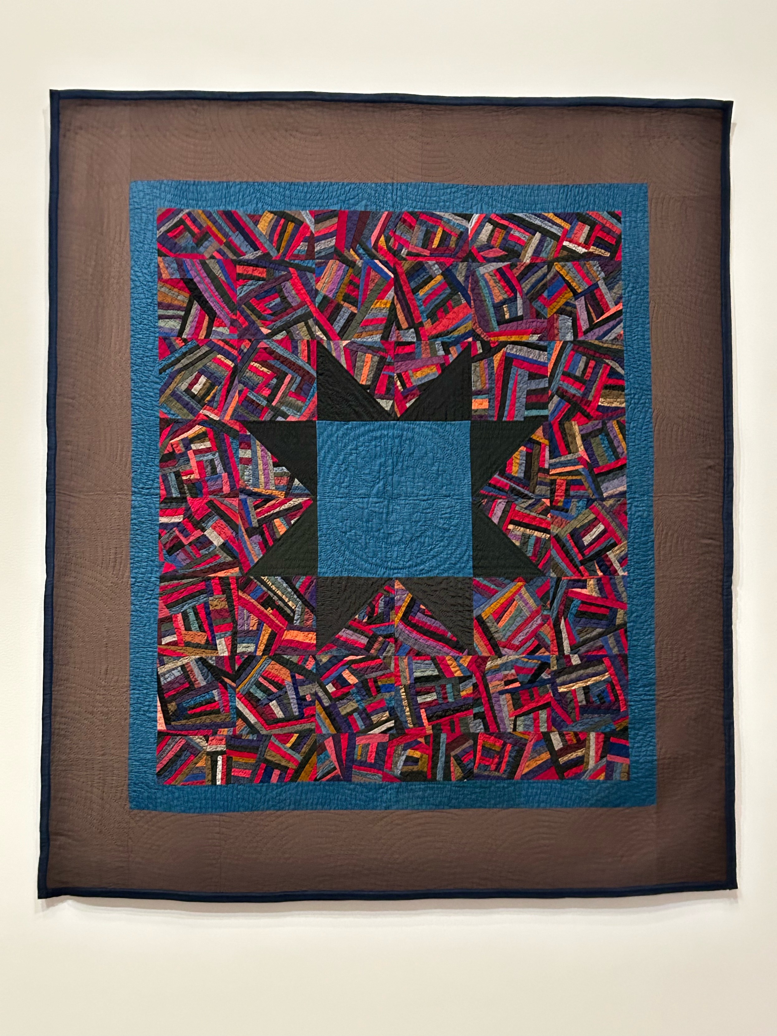 An Amish quilt with a big bold blue and black star in the center. It’s surrounded by blocks made up of tiny scraps of red and orange and black and purple and green and a little yellow. Each block is made up of multiple sections of scraps sewn together. Then there’s a thin medium blue border and a faded black border. 