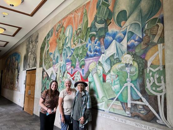 Georgia Museum of Art registrar Christy Sinksen (left), former interim director Annelies Mondi (middle) and conservator Libby Hatmaker (right) stand in front of a mural by Jean Charlot about the history of journalism, inside what was once the journalism building on the University of Georgia campus.