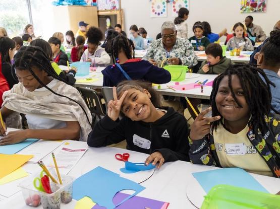 Clarke County School District 5th-graders posing for a photo in the studio classroom at the Georgia Museum of Art while they make their own abstract animal sculptures out of cut paper. The classroom is full and the kids are smiling and having a good time.