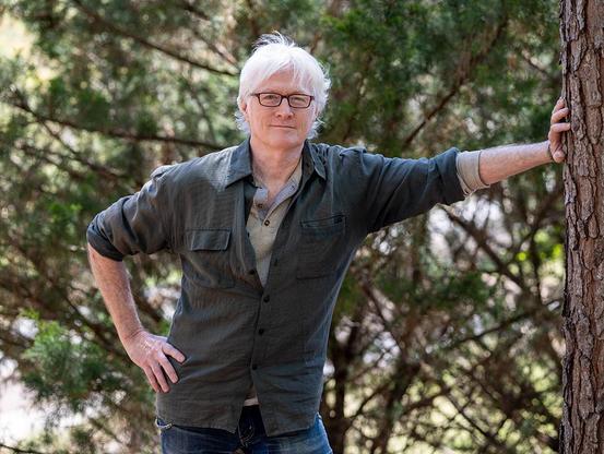 A color photograph of Doug Hollingsworth, seen about half length, with his left hand on the trunk of a pine tree (his left arm extended) and his right hand on his hip. There are more pine trees behind him.