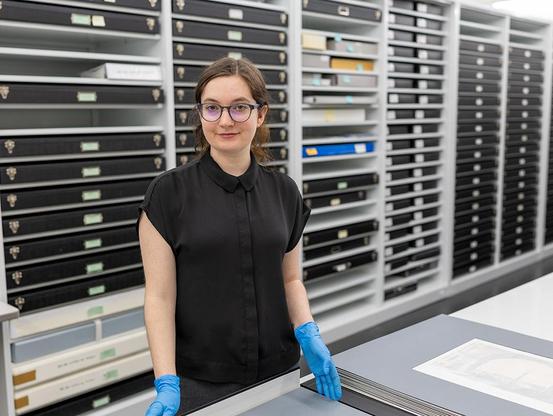 New Georgia Museum of Art assistant registrar Grace Burns poses for a photo in the works on paper vault, wearing glasses, a black top, and blue nitrile gloves.