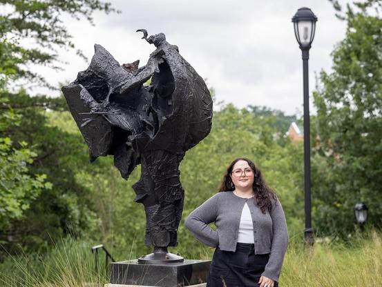 Kelsey Siegert stands to the right of a sculpture by William J. Thompson titled "Archangel." She wears a gray cardigan and stands with her right hand on her hip, near a lightpost. The scenery all around is lush and green.