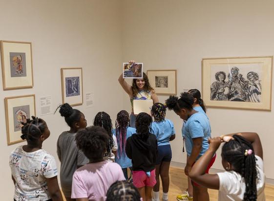 A group of children participating in an Art Adventures program at the Georgia Museum of Art. They're standing in the galleries, facing an educator who's holding up an image of a work of art.