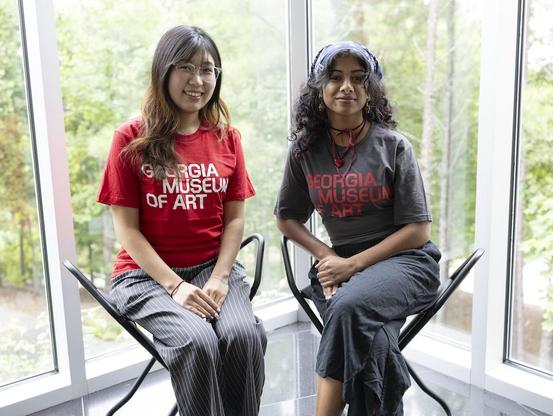 Two interns at the Georgia Museum of Art wearing t-shirts with the museum's new brand. They're sitting in gallery stools in front of a window, through which we can see trees.