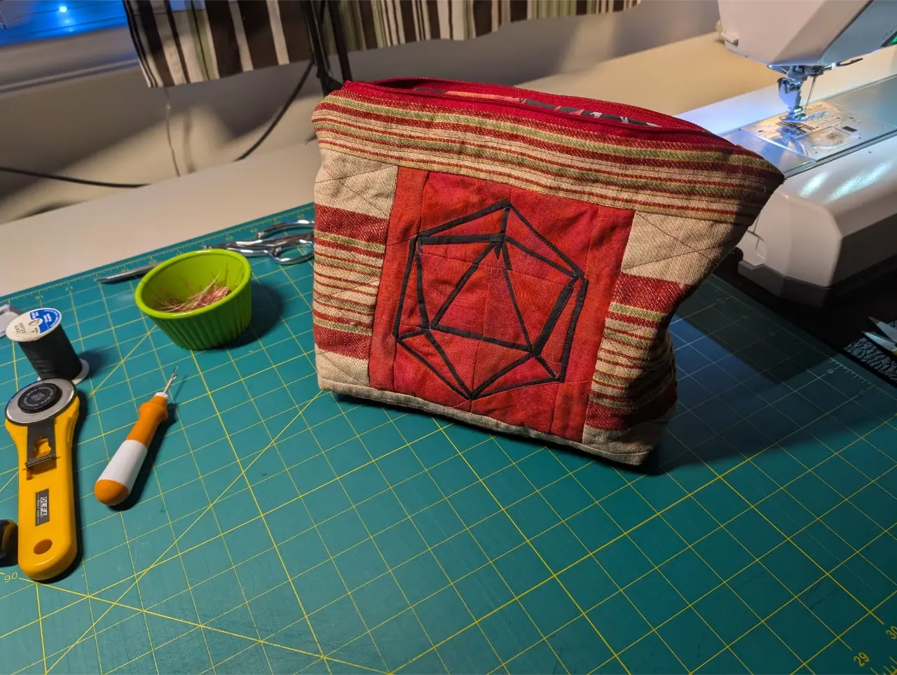 A zippered bag sits on a green cutting mat next to a sewing machine. In the center of the bag is a block with a d20 outline in black on a red background. The rest of the bag is a brown fabric with mainly red stripes.