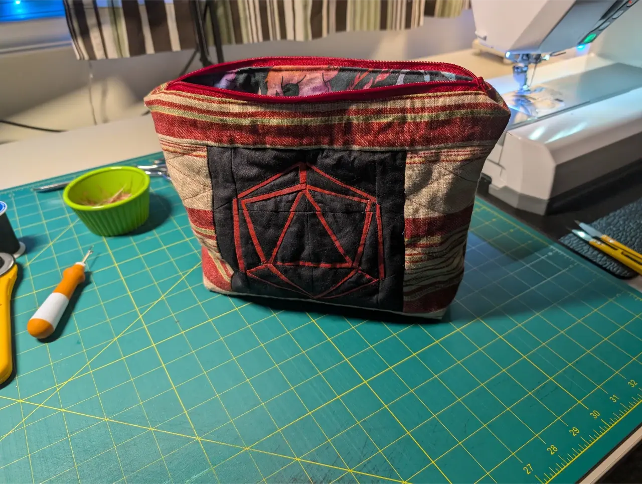 A zippered bag sits on a green cutting mat next to a sewing machine. In the center of the bag is a block with a d20 outline in red on a black background. The rest of the bag is a brown fabric with mainly red stripes.