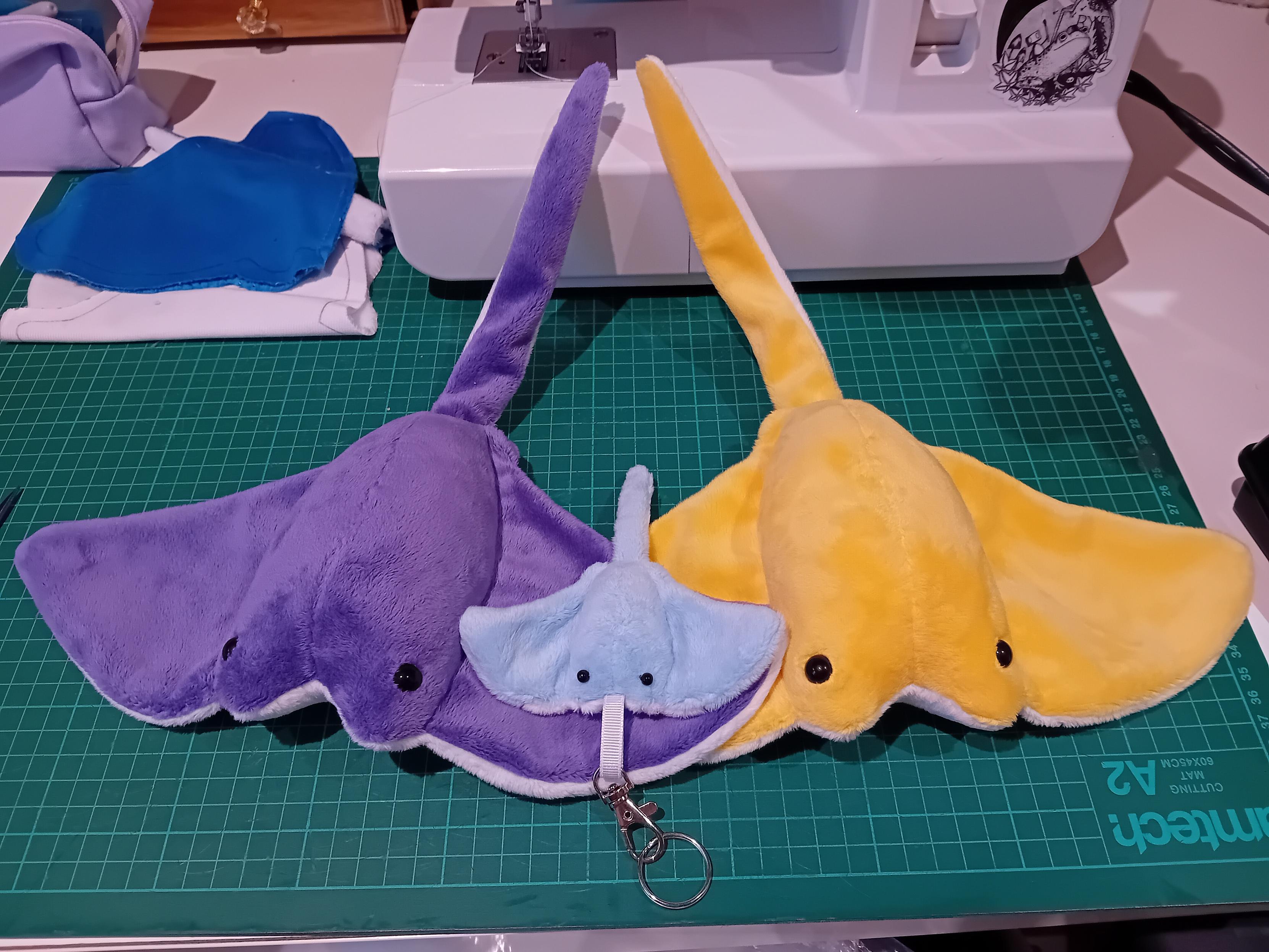 Three stingray plushies. One's purple with a white belly, another is sunshine yellow with a white belly, and nestled in amongst their wings is a much smaller, blue bag charm with a trans flag belly.