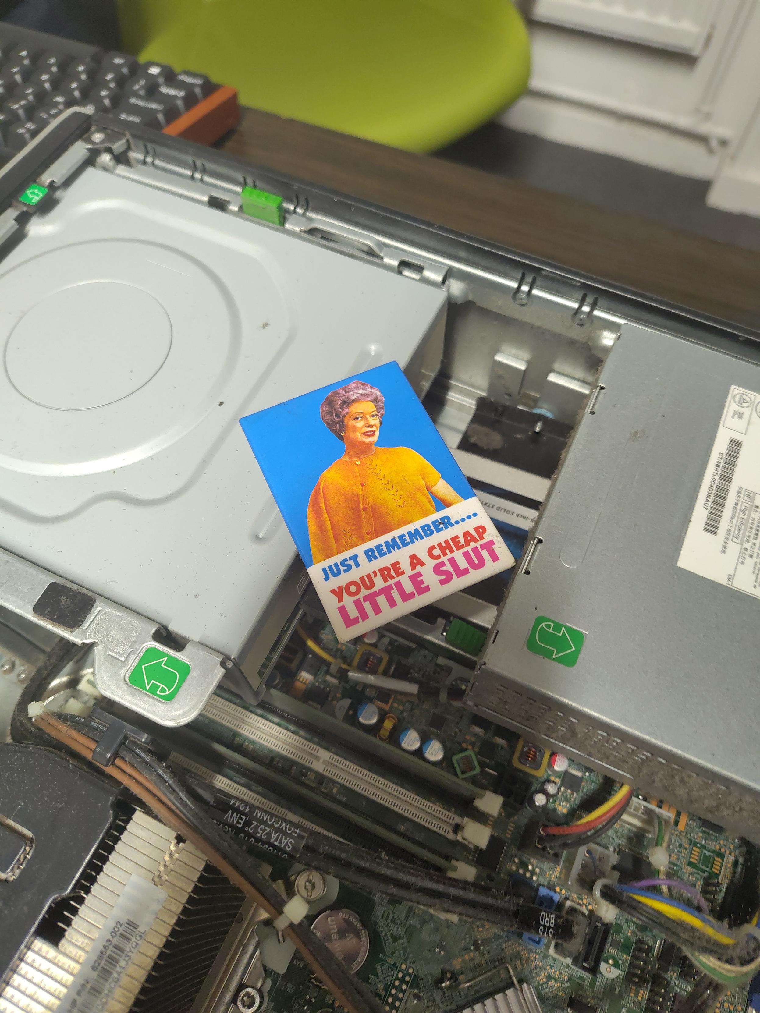 A fridge magnet with an old woman on it with the caption "Just remember... You're a cheap little slut" resting inside an opened slimline computer