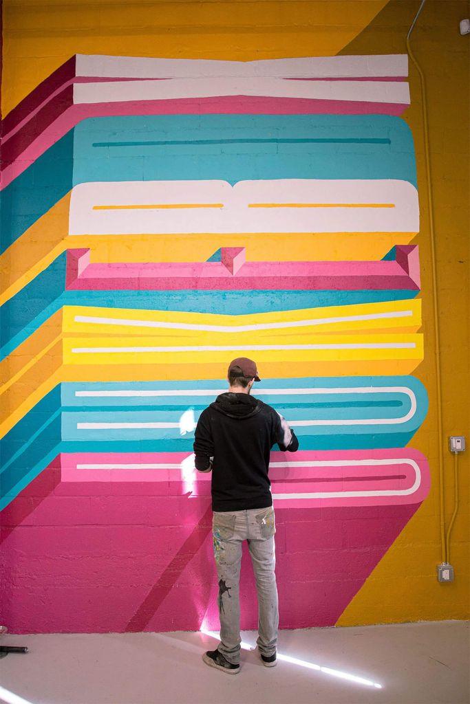 A graffiti artist in a black hoodie paints a vibrant mural of colorful geometric shapes on a yellow wall.