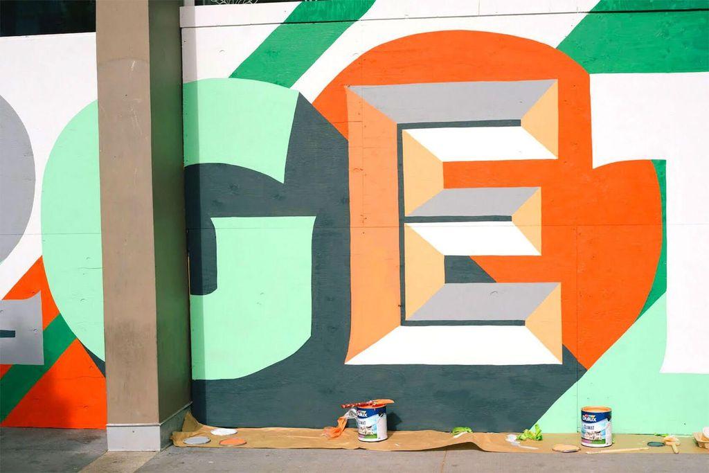 A vibrant mural featuring geometric shapes, letters and bold colors, with paint cans and tools scattered on the ground below.