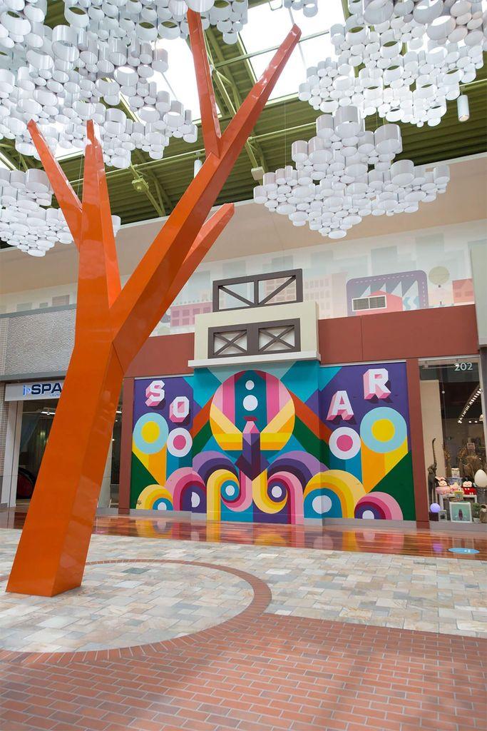 A vibrant mall interior features an orange sculptural tree and a colorful mural with geometric designs, illuminated by a modern light installation.