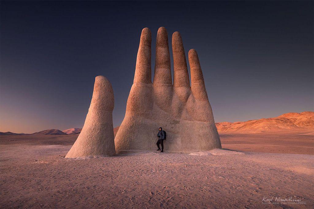 A giant hand sculpture rises from the desert landscape, with a person standing beside it against a twilight sky.