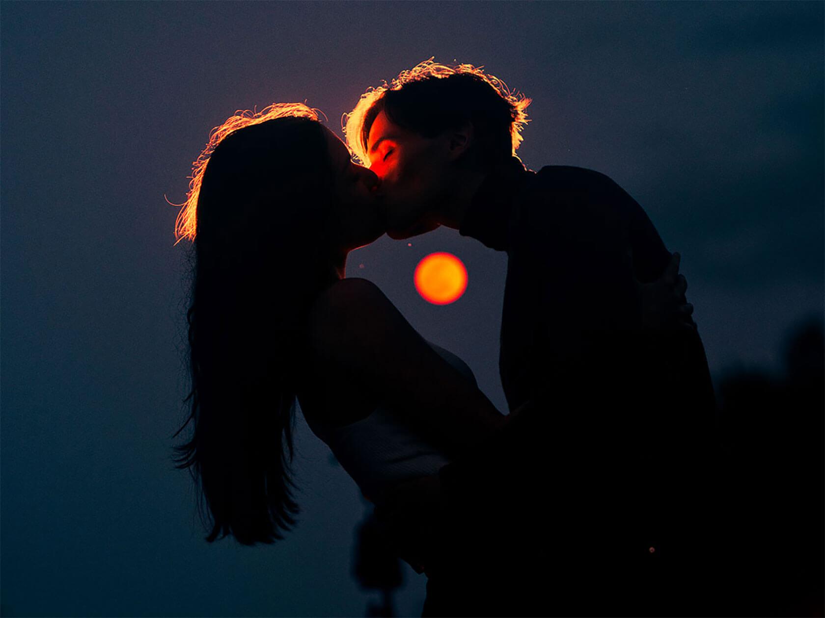Silhouetted couple kissing against a dark blue sky, their faces and hair rimmed with warm orange light and a glowing red-orange orb in the background.
