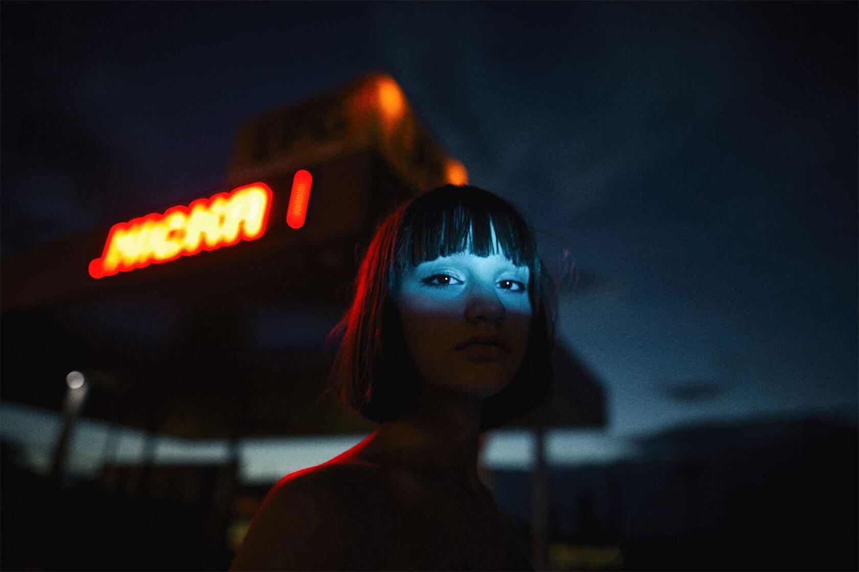 Nighttime portrait of a woman with a dark bob haircut, her eyes illuminated by a narrow beam of blue light beneath a blurred red neon sign.