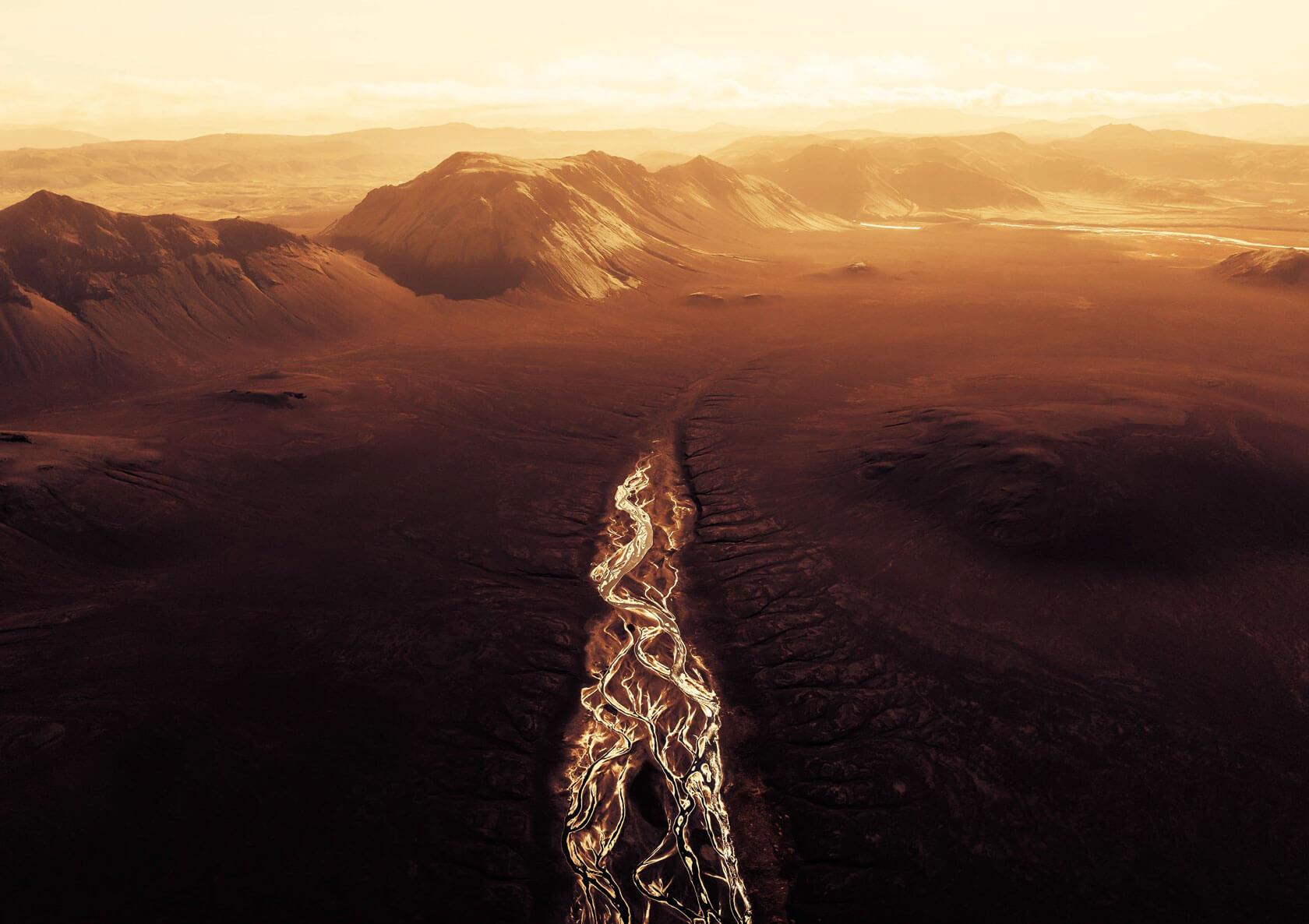 Aerial view of a braided river gleaming like molten veins across a vast rust-colored volcanic plain beneath steep, sunlit mountains and a hazy golden sky.