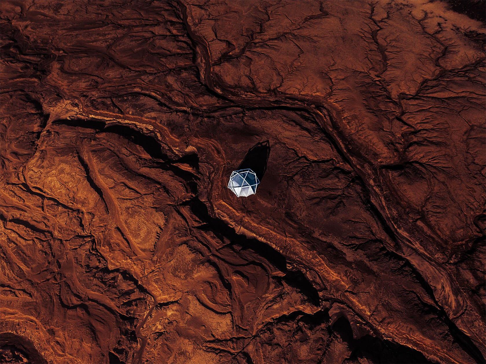 Solitary white geodesic dome casting a long shadow on a rust-red, cracked desert landscape from above, evoking an isolated research camp or Mars-like terrain.