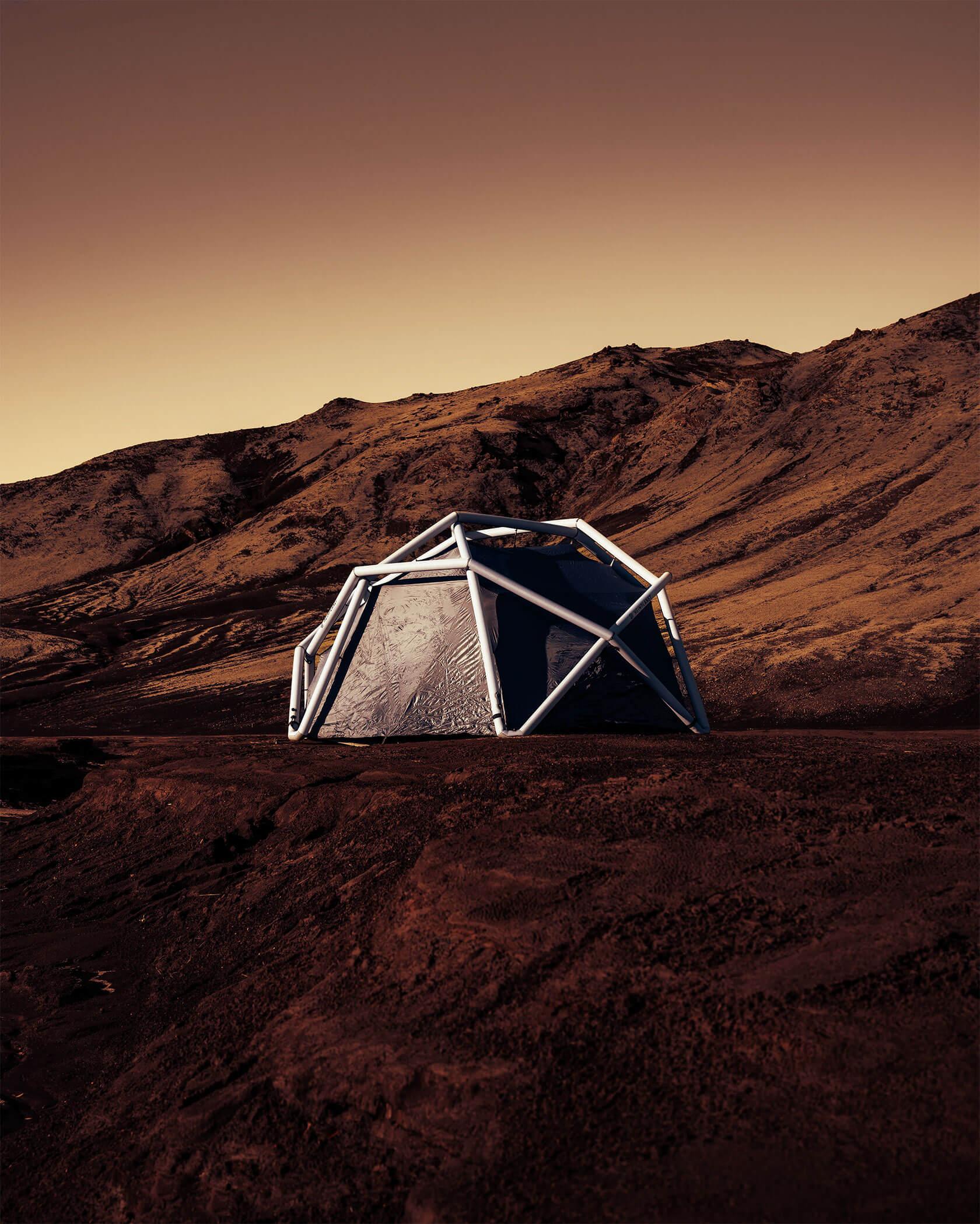 A geodesic shelter with white tubular frame pitched on reddish, rocky terrain beneath barren hills and a warm, orange sky, suggesting a remote Mars-like expedition camp.