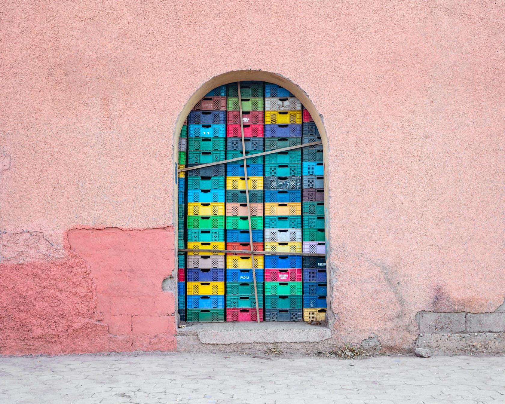 Colorful plastic crates stacked tightly inside an arched opening in a weathered pink stucco wall.