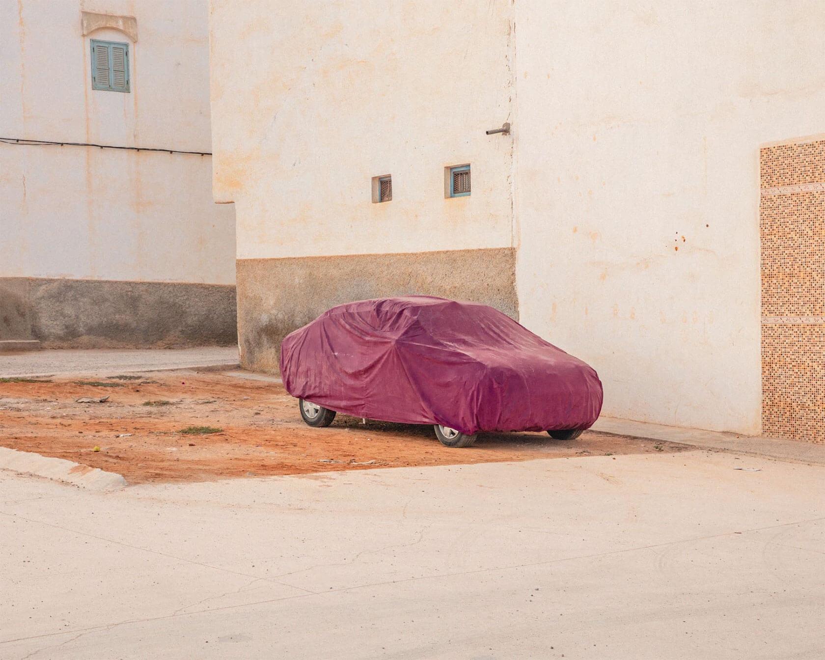 Car covered with a dusty purple fabric cover, parked beside pale stucco buildings on a quiet street.